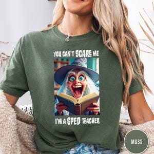 Puede incluir: Camiseta verde musgo con el texto "YOU CAN'T SCARE ME" sobre una imagen de una bruja de dibujos animados sosteniendo un libro y el texto "I'M A SPED TEACHER". La bruja tiene la boca muy abierta y lleva un sombrero.
