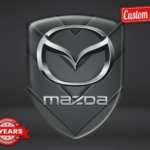 Può includere: Un emblema a forma di scudo con il logo e la scritta Mazda in argento su uno sfondo grigio scuro testurizzato. Un'etichetta rettangolare rossa nell'angolo in alto a destra riporta la scritta Custom Made e un badge circolare rosso in basso a sinistra riporta 5 YEARS WARRANTY.
