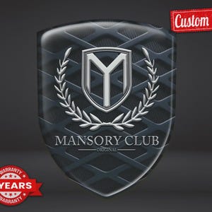 May include: A shield-shaped emblem featuring a dark grey grid pattern background. Centred is a silver crest with the letter M, surrounded by a laurel wreath. Below, the text reads MANSORY CLUB ORIGINAL. Includes a red Custom Made tag.