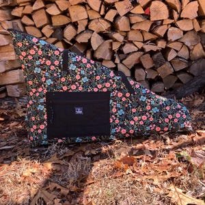 May include: A black triangular bag with a floral pattern of pink and blue flowers on a dark background. The bag has a black pocket and a handle. The bag is sitting on a bed of dry leaves.