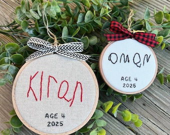 May include: Two round wooden embroidery hoop ornaments featuring white fabric. The larger ornament has red embroidered text reading Kiran and black text reading AGE 4 2025 with a black and white chevron bow. The smaller has black text reading OMON AGE 4 2025 with a red and black plaid bow.