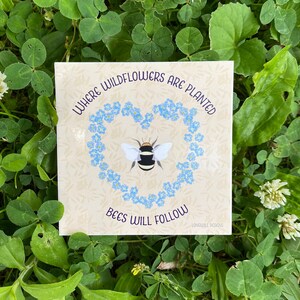 Puede incluir: Una pegatina beige con una corona de flores silvestres azules en forma de corazón que rodea a un abejorro. El texto dice "Donde se plantan las flores silvestres, las abejas seguirán". La pegatina está sobre un lecho de trébol verde.