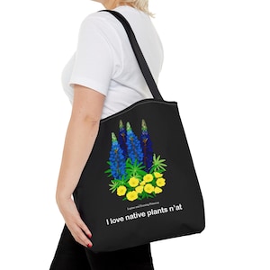 May include: Black tote bag with a design of blue and green lupine flowers and yellow evening primrose flowers. The text "I love native plants n'at" is printed on the bag.