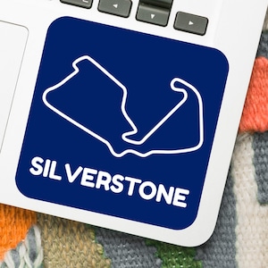 May include: A blue Silverstone sticker with a white outline of the race track, placed on a laptop. The word "SILVERSTONE" is printed in white below the track outline. A smartphone and earbuds are also visible.