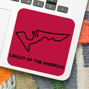 Puede incluir: Una pegatina cuadrada roja con el contorno negro del circuito "CIRCUIT OF THE AMERICAS". La pegatina está sobre un portátil, con un smartphone y auriculares cerca.