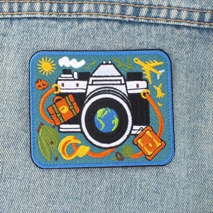 May include: A rectangular patch depicting a camera with a globe lens, surrounded by travel-related imagery such as a suitcase and aeroplane. The patch is set on a blue background and is attached to a light blue denim jacket.