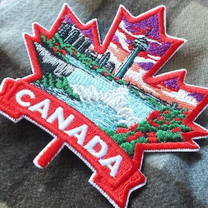 Puede incluir: Parche bordado de hoja de arce roja con la palabra "CANADA" en blanco. El parche presenta un diseño escénico de una cascada, edificios y una torre. El parche está sobre una tela a cuadros gris y marrón.