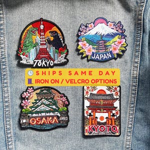 Könnte beinhalten: Vier gestickte Aufnäher mit japanischen Wahrzeichen. Die Aufnäher zeigen Tokio mit Godzilla, den Berg Fuji, die Burg Osaka und einen Tempel in Kyoto. Sie sind auf einer Jeansjacke befestigt. Die Aufnäher sind farbenfroh und detailliert.