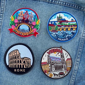 Puede incluir: Cuatro parches redondos bordados con paisajes urbanos. Los parches incluyen Ámsterdam con un puente, Praga con un castillo, Roma con el Coliseo y Viena con una catedral. Los parches están adheridos a una chaqueta vaquera.