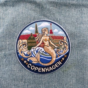 Könnte beinhalten: Runder, gestickter Aufnäher mit einer Meerjungfrau, Wellen und dem Wort "COPENHAGEN". Das Design umfasst Gebäude und einen Turm vor blau-weißem Himmel. Der Aufnäher ist auf einer Jeansjacke befestigt.