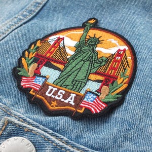 Puede incluir: Parche bordado con la Estatua de la Libertad, el Golden Gate Bridge y banderas americanas. El parche es circular con un borde negro y el texto "U.S.A." en marrón. Está cosido a una chaqueta vaquera azul claro.