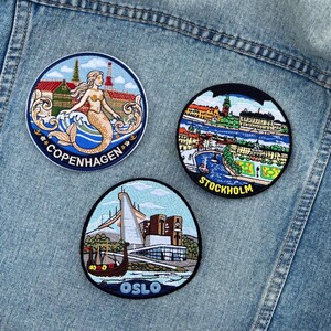 May include: Three embroidered patches on a light blue denim jacket. The patches feature cityscapes of Copenhagen, Stockholm, and Oslo. The Copenhagen patch shows a mermaid. The Stockholm patch depicts a river scene. The Oslo patch shows a building and a boat.