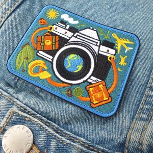 May include: Embroidered patch featuring a vintage camera design with travel-themed elements. The patch includes a globe, suitcase, airplane, and sun, all set against a teal background with a blue border. The patch is sewn onto a denim jacket.