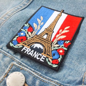 May include: Embroidered patch depicting the Eiffel Tower, the French flag, and floral details. The rectangular patch, with the word "FRANCE", is sewn onto a light blue denim jacket.