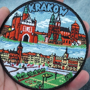 May include: Embroidered patch featuring a colorful illustration of Krakow, Poland. The design includes the city's architecture, with the word "KRAKOW" in blue letters at the top. The patch is round with a black border.