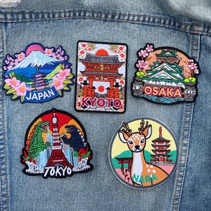 Peut inclure: Cinq écussons brodés représentant des monuments et des villes du Japon. Les motifs incluent le Mont Fuji, une pagode, un cerf et les villes de Kyoto, Osaka, Tokyo et Nara. Les écussons sont de formes et de couleurs variées.