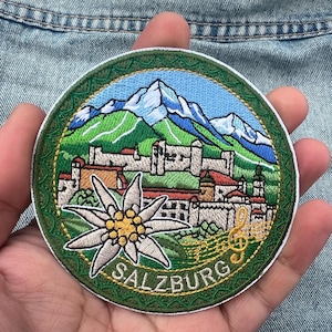 May include: Embroidered round patch featuring a scenic view of Salzburg, Austria. The design includes a mountain range, buildings, and an edelweiss flower. The word "SALZBURG" is embroidered in white. The patch is approximately 3 inches in diameter.