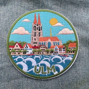 May include: Embroidered patch featuring a scenic view of Ulm, Germany. The design includes a church with red spires, buildings, water, and the word "ULM". The patch has a blue, green, and white color scheme, with a yellow sun and fluffy clouds.