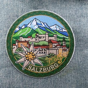 May include: Embroidered patch featuring a scenic view of Salzburg, Austria. The design includes a mountain range with snow-capped peaks, a cityscape with buildings, and an edelweiss flower. The word "SALZBURG" is arched along the bottom edge in a gold color.