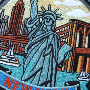 May include: Embroidered patch featuring the Statue of Liberty, New York City skyline, and Brooklyn Bridge. The patch has a blue banner with the text "NEW YORK" in red. The design includes boats and clouds.
