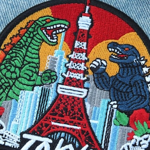 May include: Embroidered patch featuring two Godzilla-like monsters facing each other near the Tokyo Tower. The patch has a black border, with the word "Tokyo" at the bottom. The background includes a red sky and a cityscape.