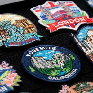 May include: A collection of embroidered travel patches featuring iconic landmarks including the Eiffel Tower labelled France, the Statue of Liberty labelled New York, a London scene, the Taj Mahal, Mount Fuji labelled Japan, and Yosemite California.