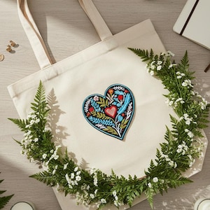 May include: Cream-colored tote bag with a heart-shaped floral design. The heart features red, blue, and green details. The bag is surrounded by a wreath of greenery and small white flowers. A gold necklace and earrings are visible.