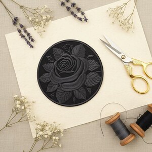 Puede incluir: Un parche de rosa negra bordada sobre papel crema, con tijeras doradas y dos carretes de hilo. Lavanda seca y flores blancas están dispuestas alrededor del parche. El parche presenta un diseño de rosa detallado.