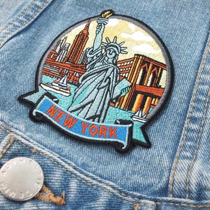 Puede incluir: Parche bordado de la ciudad de Nueva York con la Estatua de la Libertad, rascacielos y veleros. El parche tiene una pancarta azul con las palabras "NEW YORK" en letras naranjas. El parche está cosido a una chaqueta vaquera azul claro.