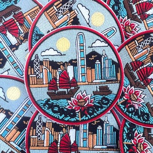 Peut inclure: Écussons brodés représentant un paysage urbain avec des bateaux, un soleil et une fleur de lotus. Les écussons ont une bordure rouge et un fond bleu clair. Le mot "HONG KONG" est brodé en bas de l'écusson.