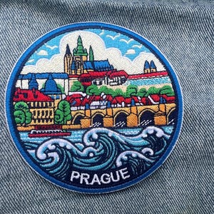 Peut inclure: Écusson brodé représentant Prague, République tchèque, avec des bâtiments, un pont et de l'eau. Le nom de la ville est en texte blanc. L'écusson a une bordure bleue et est posé sur un fond en denim.
