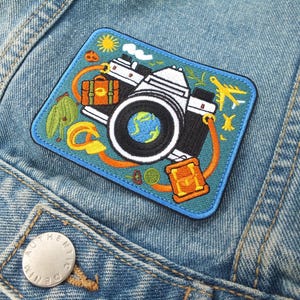 May include: Embroidered patch featuring a vintage camera design with a globe in the lens. The patch includes travel-themed elements like a suitcase, airplane, and sun, set against a blue background. It is sewn onto a denim jacket.