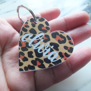 Puede incluir: Llavero en forma de corazón con estampado de leopardo en beige, negro y naranja. El nombre "olivia" está escrito en escritura blanca en el centro. Un llavero de metal plateado está adjunto en la parte superior.