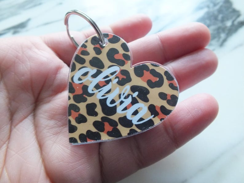 Puede incluir: Llavero en forma de coraz&oacute;n con estampado de leopardo en beige, negro y naranja. El nombre "olivia" est&aacute; escrito en escritura blanca en el centro. Un llavero de metal plateado est&aacute; adjunto en la parte superior.