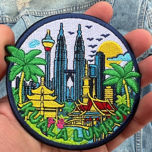 May include: Embroidered patch featuring a colorful cityscape of Kuala Lumpur, Malaysia. The design includes the Petronas Towers, palm trees, and a setting sun. The word "KUALA LUMPUR" is embroidered in green.