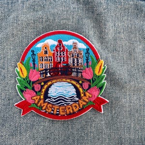 May include: Embroidered patch featuring a colorful Amsterdam scene. The design includes iconic canal houses, a bridge, tulips, and the word "AMSTERDAM" in orange. The patch is set against a denim background.
