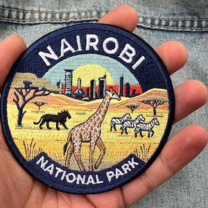 May include: A circular embroidered patch featuring a savanna landscape with a giraffe, a black lion, and two zebras. The background shows a yellow sun, a city skyline, and acacia trees. The dark blue border contains the white text NAIROBI NATIONAL PARK.