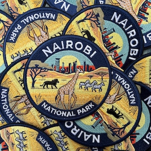 May include: A collection of circular embroidered patches featuring a landscape of Nairobi National Park. Each patch displays a giraffe, zebras, and a lion silhouette against a yellow savanna, with a city skyline and the text NAIROBI NATIONAL PARK.