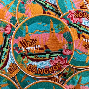 Könnte beinhalten: Runde, gestickte Aufnäher mit einer farbenfrohen Darstellung eines thailändischen Tempels und dem Schriftzug "Bangkok". Die Aufnäher haben einen türkisfarbenen Hintergrund mit orangefarbenen, rosa und grünen Details. Die Aufnäher haben einen weißen Rand.