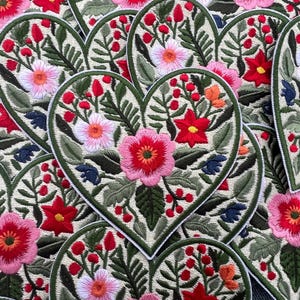 Op de afbeelding: Geborduurde hartvormige patches met bloemmotieven. Elke patch heeft een crèmekleurige achtergrond met een groene rand en is gevuld met roze, rode en witte bloemen, groene bladeren en rode bessen. Een verzameling decoratieve patches.