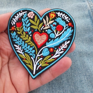 May include: Embroidered heart-shaped patch with a black background. The patch features a central red heart, surrounded by blue, green, and white floral and leaf designs. The patch is held in a hand.