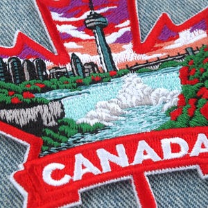 Puede incluir: Parche bordado con forma de hoja de arce roja con una detallada representación de un paisaje canadiense. El diseño incluye una cascada, un paisaje urbano y la Torre CN. La palabra "CANADA" está bordada en blanco.
