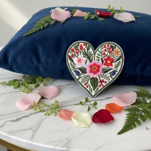 Op de afbeelding: Een hartvormige geborduurde patch met een bloemenontwerp in roze, rode en oranje tinten, omgeven door groene bladeren. De patch ligt op een donkerblauw fluwelen kussen, met verspreide bloemblaadjes.