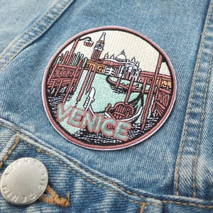May include: Embroidered patch depicting Venice, Italy, with a gondola, buildings, and the word "VENICE" in teal. The round patch, with a pink border, is sewn onto a light blue denim jacket.