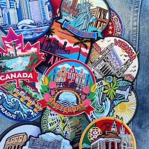 May include: A collection of embroidered travel patches featuring iconic landmarks and cityscapes. Patches include New York, Canada, Amsterdam, Vienna, and Prague. The patches are in various shapes and colors, with detailed designs.