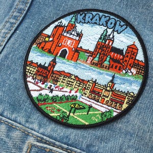 May include: Embroidered patch featuring a colorful illustration of Krakow, Poland, with the city's name in blue letters. The patch depicts buildings, a river, and green spaces, set against a blue sky. The patch is circular with a black border, sewn onto a denim jacket.