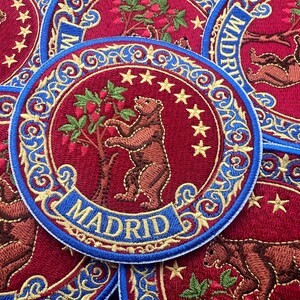 May include: Embroidered patches with the Madrid coat of arms. The design shows a brown bear and a strawberry tree, surrounded by stars and ornate blue and gold trim. The word "MADRID" is displayed in a banner.