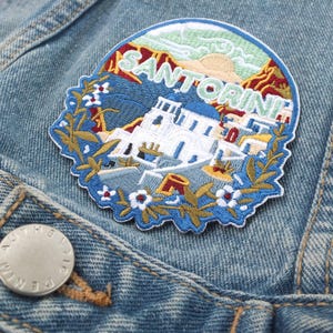 May include: Embroidered patch featuring a blue, white and yellow image of a town on a hillside with the text "SANTORINI" in white. The patch is surrounded by a green wreath with white flowers.