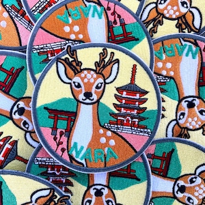 Peut inclure: Écussons brodés avec un cerf, une pagode japonaise et le mot "NARA" en turquoise. Les écussons circulaires ont une bordure grise. Le fond est bleu clair.