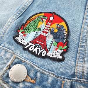 May include: Embroidered patch depicting Godzilla monsters, the Tokyo Tower, and the word "TOKYO". The patch is on a light blue denim jacket. The patch has a black border and a red, green, and blue colour scheme.